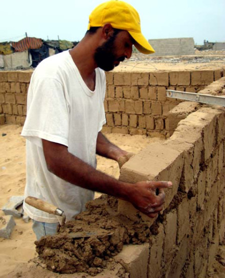 A man building a wall of bricks. The wall is chest-high to the man so far, he is using a trowel with a bucket of mortar to add new bricks.