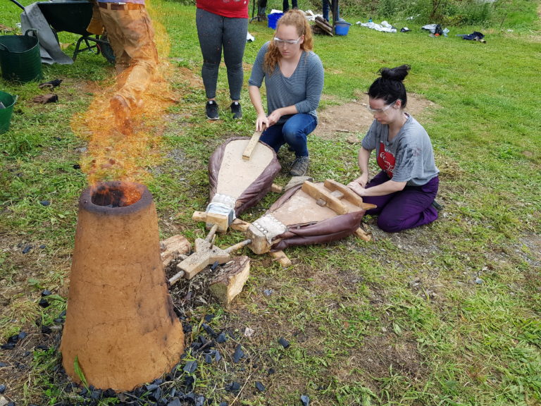 A simple cylindrical clay furnace, with flames coming out of the top. Two young women are kneeling in front of it, pushing on bellows (air pumps) to drive air through it.