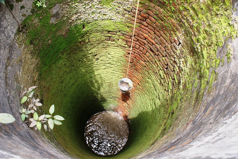 A photo looking into a well lined with bricks, a bucket hanging down on a rope, with water at the bottom.