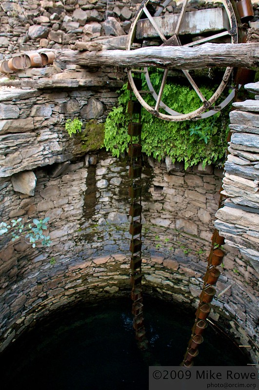 View inside a well with walls of stone and mortar. There is a bucket chain: a chain of small metal buckets looped over a big wooden wheel, so that as you pull the chain the rising buckets automatically bring up water.