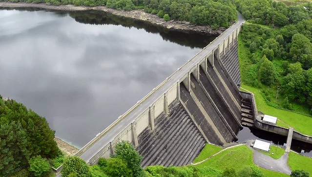 Photo of a reservoir: it looks like an long, thin lake, flat water surrounded by greenery. At one end there is an escarpment where the ground drops back steeply and the water is held back by a concrete barrier with 'gates' that lead to a narrow concrete-lined channel of water flowing downhill.